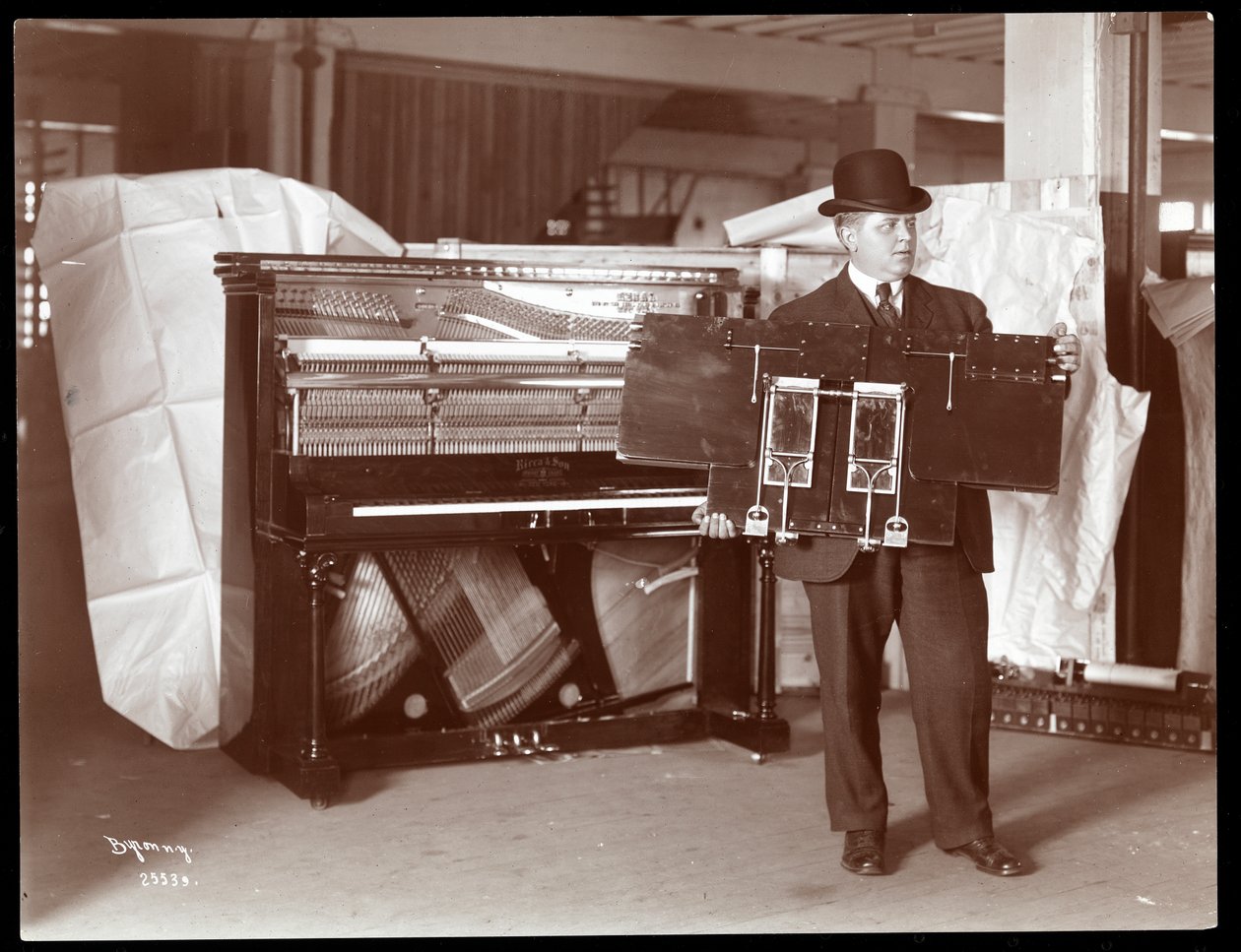 بيانو لشركة Ricca und Son Piano Co مع رجل مجاور يحمل جزءًا من أعمال البيانو ، 1907 (طباعة الجيلاتين الفضي) بواسطة شركة بايرون