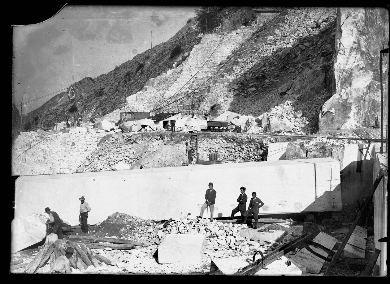 ثلاثة رجال يراقبون عمل العديد من الحفارات في Apuan Alps. خلفهم كتلة ضخمة من الرخام مربعة بالفعل وجاهزة للنقل إلى الوادي (صورة بالأبيض والأسود) بواسطة أ. كورسيني
