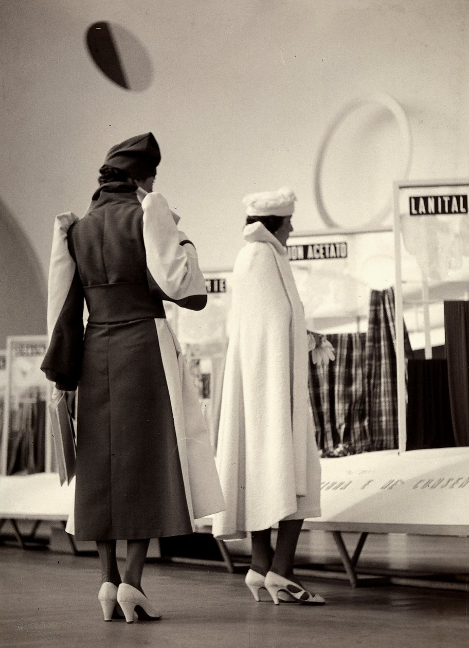 نموذجان من الخلف يرتديان إبداعات دار الأزياء الراقية "لما". ترتدي إحدى العارضات معطفاً ذا لونين مُجمَّع مع وشاح عند الخصر. النموذج الآخر يرتدي عباءة كاملة. القبعات من نفس اللون تكمل كل مجموعة. (ب / ث الصورة) بواسطة لويس بوجينو