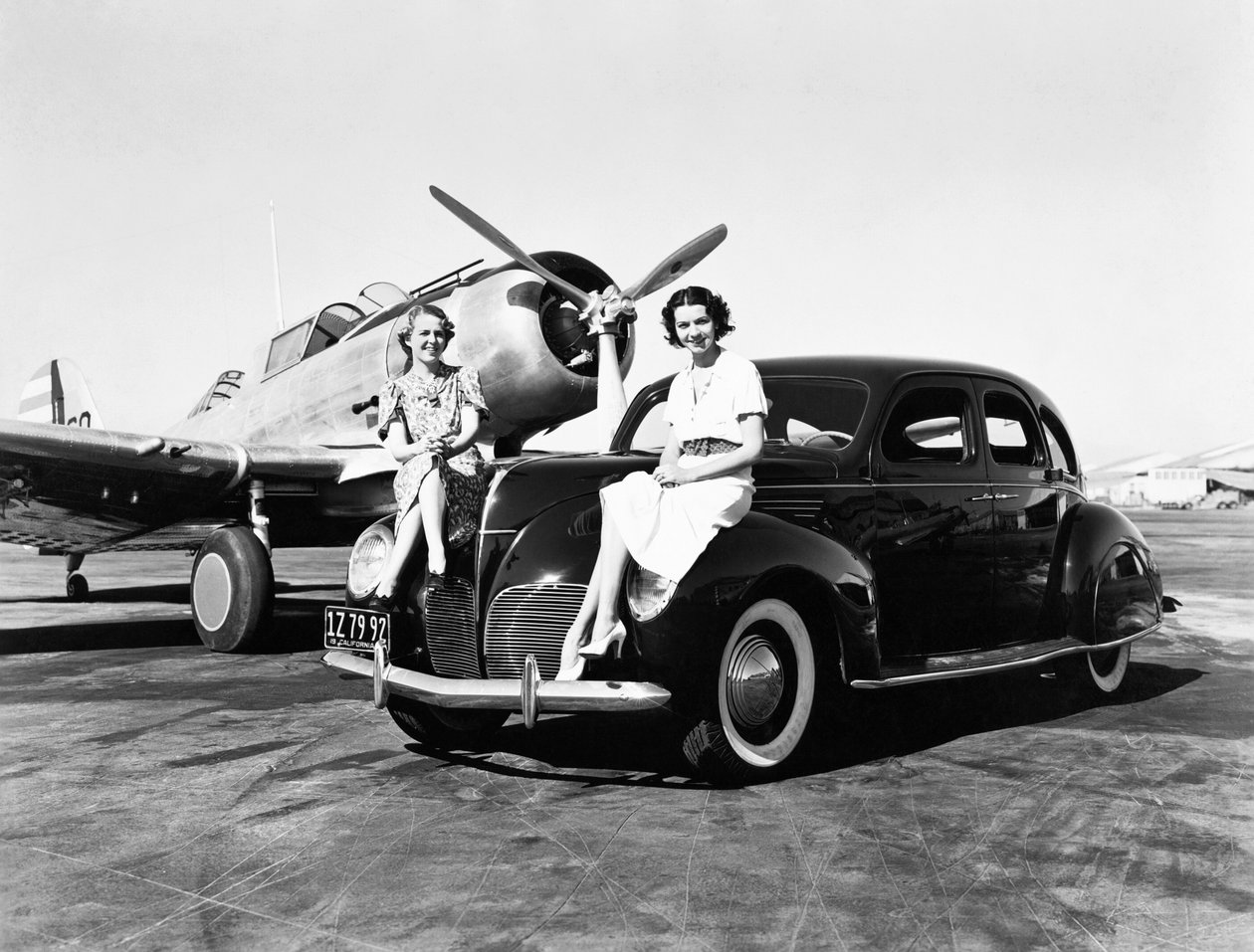 كاليفورنيا: 1939 امرأتان تجلسان على مصدات سيارة لينكولن زفير سيدان عام 1939 في مطار مع طائرة ذات محرك واحد متوقفة خلفهما (صورة أبيض وأسود) بواسطة غير معروف
