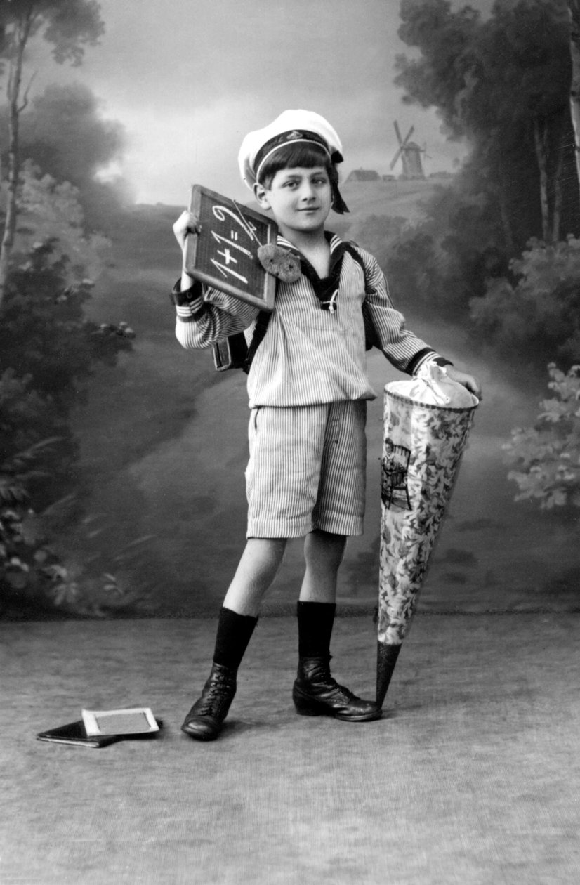 First day of school. A young boy in uniform posing with a slate showing the addition 1+1 = 2 and a surprise pouch. Photography around 1910. بواسطة فرديناند جورج والدمولر
